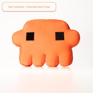 Peut inclure: Un coussin en peluche orange vif en forme de créature pixélisée avec deux yeux carrés noirs. Au-dessus du coussin, une étiquette rectangulaire à bordure orange affiche le texte : Set 1 sold out - Preorder Set 2 now.