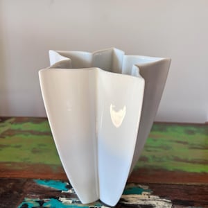 May include: A white ceramic vase with a unique, star-like shape. The vase has multiple triangular panels that create a textured, geometric design. The glossy finish reflects light, highlighting the vase's form.