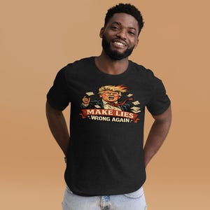 Może przedstawiać: Czarny t-shirt z kreskówkową ilustracją mężczyzny z blond włosami i czerwoną krawatem. Grafika zawiera napis "MAKE LIES WRONG AGAIN" na czerwonym banerze. Projekt jest wyśrodkowany na przodzie koszulki.