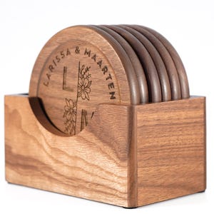 Op de afbeelding: Een set van zes ronde houten onderzetters met een bijpassende houder. De onderzetters zijn gegraveerd met de namen "LARISSA & MAARTEN" en bloemmotieven. De houder is een rechthoekige doos met een gebogen uitsparing.