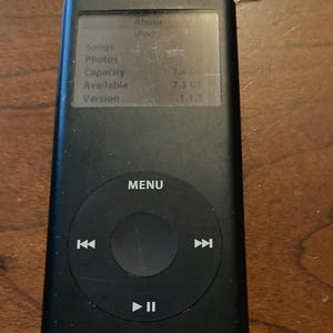 Könnte beinhalten: Schwarzer iPod Digital-Media-Player mit einem Bildschirm, der Geräteinformationen anzeigt. Das Gerät hat ein kreisförmiges Bedienfeld mit einer Menütaste und Wiedergabesteuerungen. Der Bildschirm zeigt "Songs", "Photos", "Capacity", "Available" und "Version".