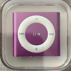 Pode incluir: Um iPod Shuffle roxo em uma caixa de plástico transparente. O dispositivo possui um painel de controle circular branco com botões de reproduzir/pausar, pular e volume. O iPod tem um design pequeno e quadrado.