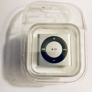 Pode incluir: Um Apple iPod Shuffle prateado em sua embalagem original. O iPod tem um botão circular preto com botões de reprodução, pausa e avanço.