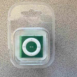 Puede incluir: Un pequeño reproductor de música digital verde con una rueda de control blanca y botones de reproducción/pausa, encerrado en un embalaje de plástico transparente. El dispositivo es rectangular con esquinas redondeadas. El paquete tiene un agujero para colgar en la parte superior.
