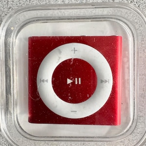 Könnte beinhalten: Ein roter iPod Shuffle in einer transparenten Kunststoffhülle. Das Gerät hat einen weißen Steuerring mit Wiedergabe/Pause-, Überspringen- und Lautstärketasten. Der iPod ist quadratisch und scheint ein Vintage-Modell zu sein.