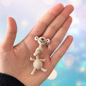 Puede incluir: Pequeña figura de perezoso de ganchillo beige con detalles blancos. El perezoso tiene ojos grandes con bordes blancos y un cuello anudado. Sus brazos y piernas están extendidos. La figura se sostiene en una mano abierta.
