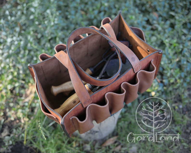 May include: A brown leather tool bag with multiple pockets and a handle. The bag is open and filled with gardening tools.