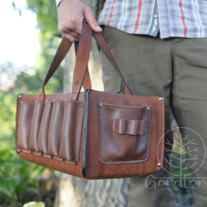 May include: A brown leather bag with multiple pockets and a handle. The bag has a flap with a small pocket on the front. The bag is being held by a person.