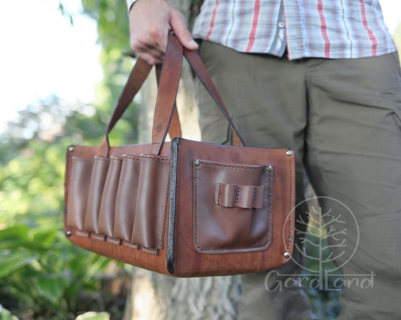 May include: A brown leather bag with multiple pockets and a handle. The bag has a flap with a small pocket on the front. The bag is being held by a person.