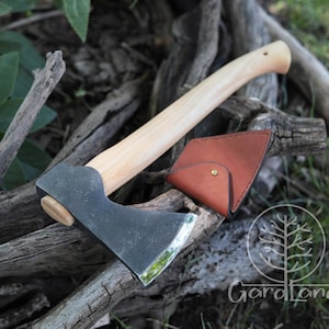May include: A black axe with a wooden handle and a brown leather sheath. The axe is resting on a pile of branches.