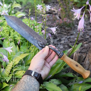 May include: A hand holds a forged gardening tool with a wooden handle. The tool has a leaf-shaped detail on the blade and a logo on the handle. The tool is surrounded by green foliage and purple flowers.