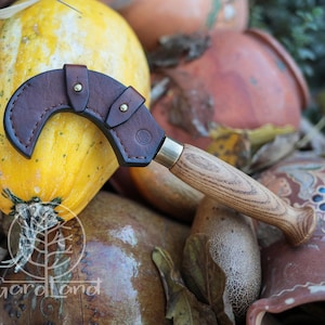 Puede incluir: Un cuchillo curvo con mango de madera y funda de cuero marr&oacute;n. El cuchillo descansa sobre una calabaza amarilla, con otras calabazas y cer&aacute;mica en el fondo. El cuchillo tiene una virola de lat&oacute;n.