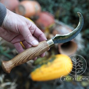 Puede incluir: Un cuchillo de hoja curva con mango de madera y detalles de lat&oacute;n. La hoja tiene un acabado oscuro y una forma curva. El mango es de madera clara con un patr&oacute;n de vetas. El cuchillo se sostiene en una mano.