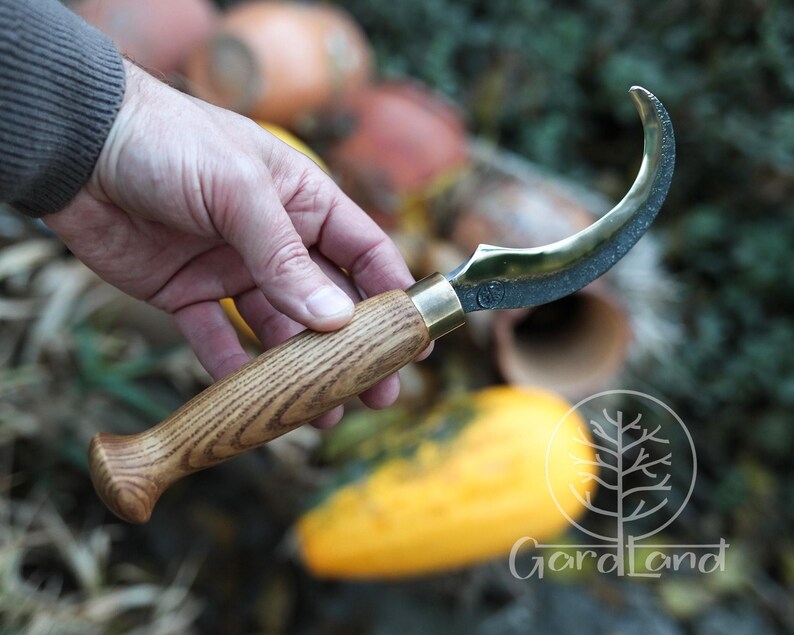 Puede incluir: Un cuchillo de hoja curva con mango de madera y detalles de lat&oacute;n. La hoja tiene un acabado oscuro y una forma curva. El mango es de madera clara con un patr&oacute;n de vetas. El cuchillo se sostiene en una mano.