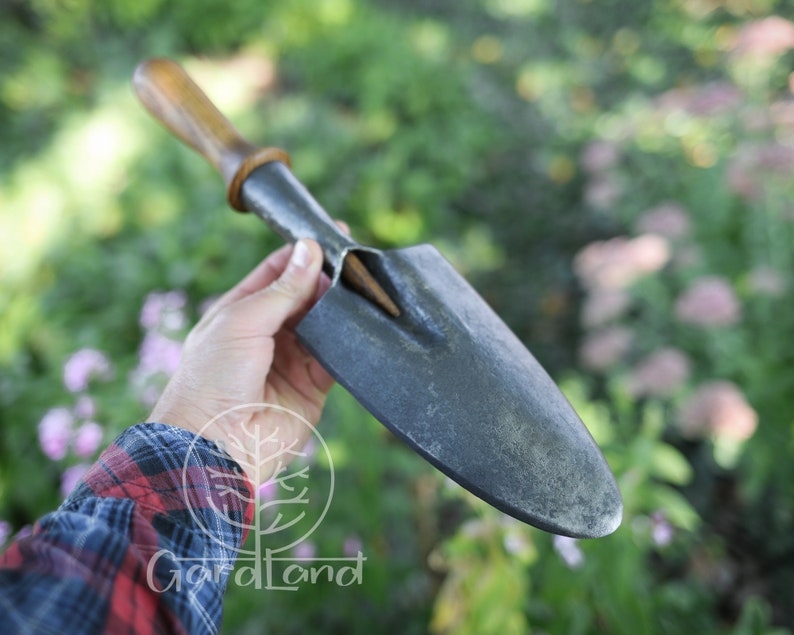 May include: A hand holds a gardening tool with a wooden handle and a metal blade. The blade is slightly curved and has a pointed tip. The tool is used for digging and planting.