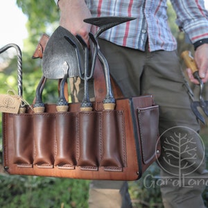 May include: A brown leather tool belt with multiple pockets for holding gardening tools. The belt is filled with various gardening tools, including a trowel, a hand rake, a cultivator, and a pruning shears. The belt is labeled "Gardaland".