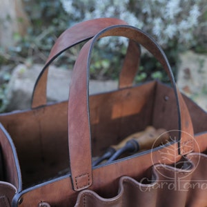 May include: A brown leather gardening tool tote with multiple pockets and a sturdy handle. The tote is empty and ready to be filled with gardening tools.