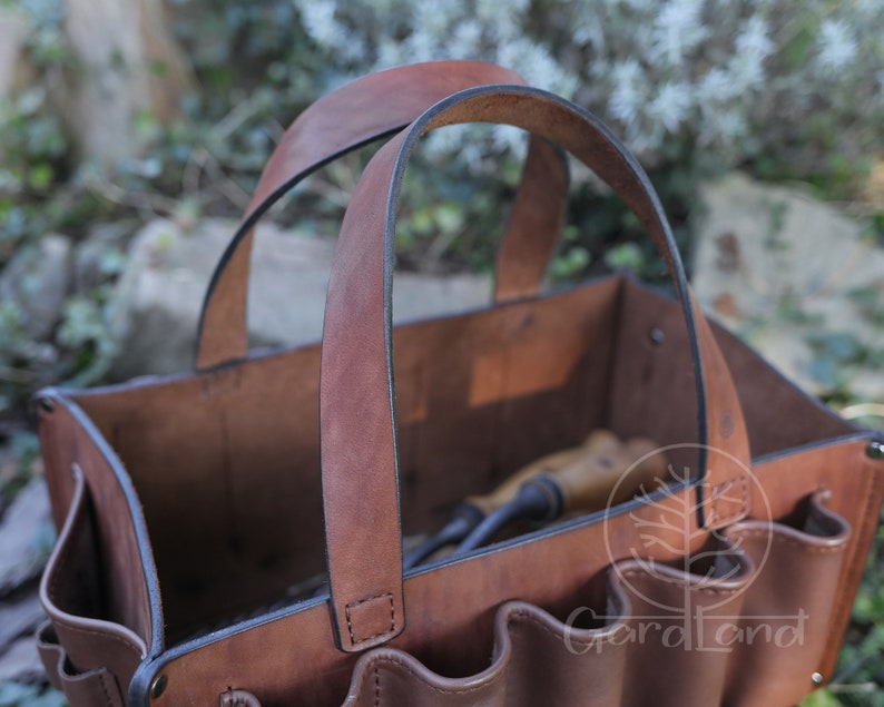 May include: A brown leather gardening tool tote with multiple pockets and a sturdy handle. The tote is empty and ready to be filled with gardening tools.