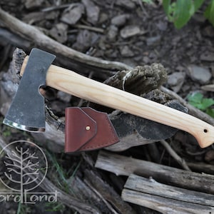 Könnte beinhalten: Eine schwarze Axt mit einem Holzstiel und einer braunen Lederscheide. Die Axt liegt auf einem Stück Holz.