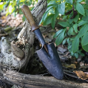 May include: A small hand forged garden trowel with a wooden handle. The trowel is black and has a slightly curved blade. The handle is brown and has a natural finish.
