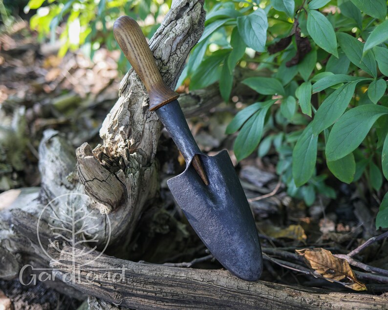 May include: A small hand forged garden trowel with a wooden handle. The trowel is black and has a slightly curved blade. The handle is brown and has a natural finish.
