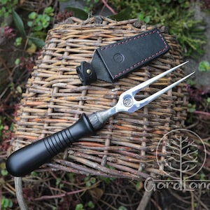 Puede incluir: Un tenedor para trinchar con mango negro y dos púas, apoyado sobre una cesta de mimbre. Una funda de cuero negro con costuras rosas está al lado del tenedor. El tenedor tiene una cabeza de metal plateado con un emblema decorativo.