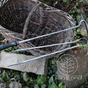 Può includere: Un rustico cesto di vimini intrecciato con manico curvo, accompagnato da tre attrezzi da giardinaggio in metallo. Gli attrezzi hanno steli in metallo ritorto e manici neri. Gli attrezzi sono appoggiati su una superficie di pietra con vegetazione.