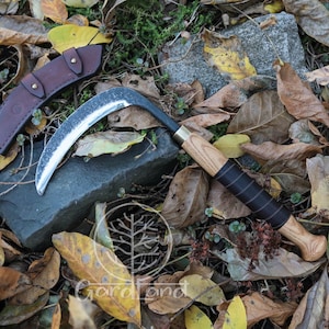 May include: A curved blade sickle with a wooden handle and black grip, resting on a stone among fallen leaves. A brown leather sheath is positioned nearby. The sickle has a silver blade and a black curved handle.