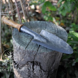 May include: A small, hand-forged garden trowel with a wooden handle. The trowel is resting on a tree stump.