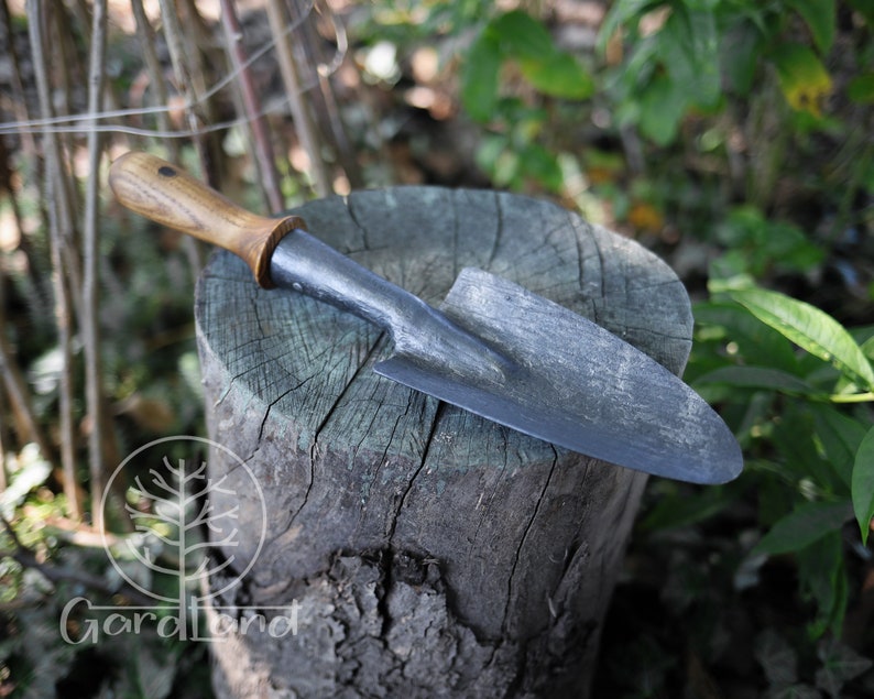 May include: A small, hand-forged garden trowel with a wooden handle. The trowel is resting on a tree stump.