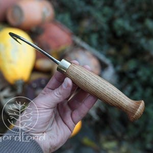 Puede incluir: Una herramienta de jard&iacute;n artesanal con un mango de madera y un gancho de metal curvado. El mango es de madera marr&oacute;n claro con una f&eacute;rula de lat&oacute;n. El gancho es negro y parece afilado. La herramienta est&aacute; dise&ntilde;ada para tareas de jardiner&iacute;a.