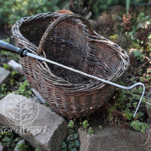 Può includere: Un cesto di vimini marrone con manico, accanto a un attrezzo da giardinaggio con manico nero ed estremità metallica curva. L'attrezzo è appoggiato sul cesto. La scena è ambientata all'aperto tra rocce e vegetazione.