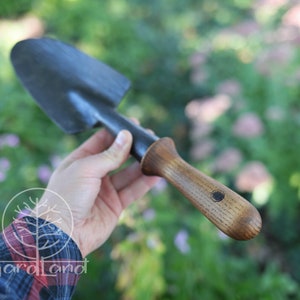 May include: A black metal garden trowel with a wooden handle. The handle has a dark brown stain and a small circular logo.