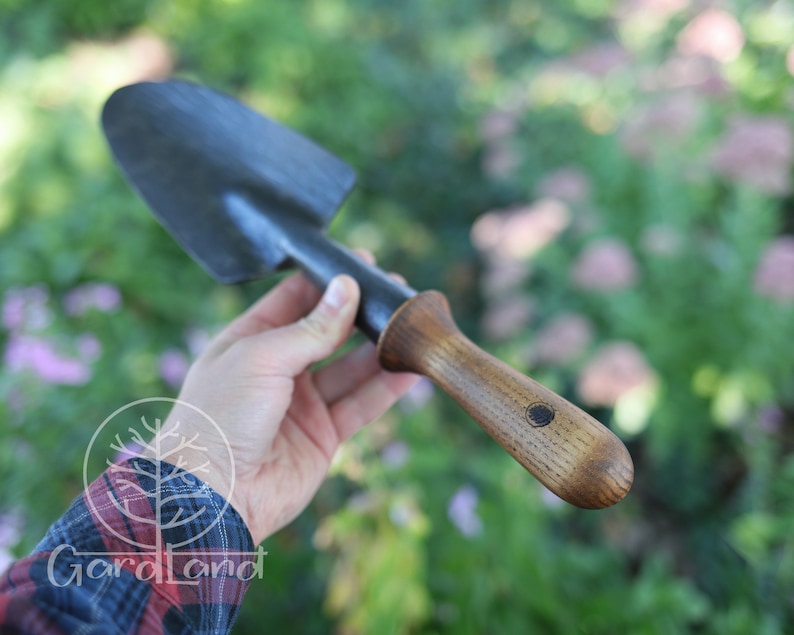 May include: A black metal garden trowel with a wooden handle. The handle has a dark brown stain and a small circular logo.