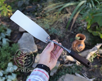 May include: A hand holds a long, thin, metal spatula with a dark wood handle. The spatula has a twisted metal handle and a flat, rectangular blade. The blade is made of stainless steel and has a copper rivet at the top.