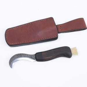 May include: A curved, stainless steel pruning knife with a dark wood handle and a small brush attached. The knife is in a brown leather sheath with a black strap.