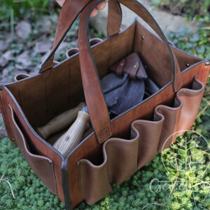 May include: A brown leather gardening tool bag with multiple pockets and a handle. The bag is filled with gardening tools, including a trowel, a hand rake, and a pair of pruning shears.