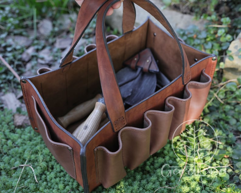 May include: A brown leather gardening tool bag with multiple pockets and a handle. The bag is filled with gardening tools, including a trowel, a hand rake, and a pair of pruning shears.