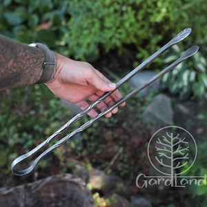 Puede incluir: Pinzas de metal forjadas a mano con diseño de hojas en las puntas y mango retorcido. Las pinzas son de color gris oscuro y se sostienen sobre un fondo de follaje verde. El logotipo de GardLand es visible en la esquina inferior derecha.