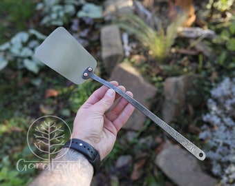May include: A hand holds a long, thin, silver spatula with a flat, rectangular head. The spatula has a hammered finish and a small hole at the end of the handle.