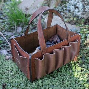 May include: A brown leather tool bag with multiple pockets and a handle. The bag is sitting on a bed of green moss.