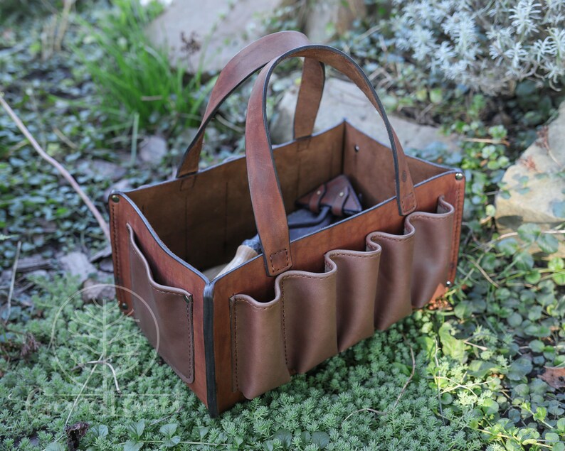 May include: A brown leather tool bag with multiple pockets and a handle. The bag is sitting on a bed of green moss.