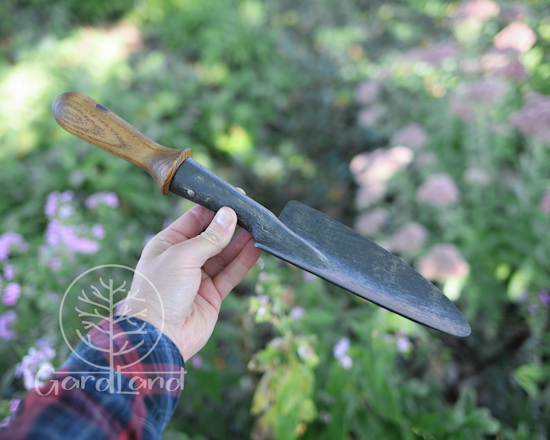 May include: A hand holds a gardening tool with a wooden handle and a black metal blade. The blade is slightly curved and has a pointed tip. The tool is likely used for digging or weeding.