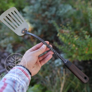 May include: A hand holds a metal spatula with a twisted handle and a wooden handle. The spatula has a slotted head and is made of stainless steel.