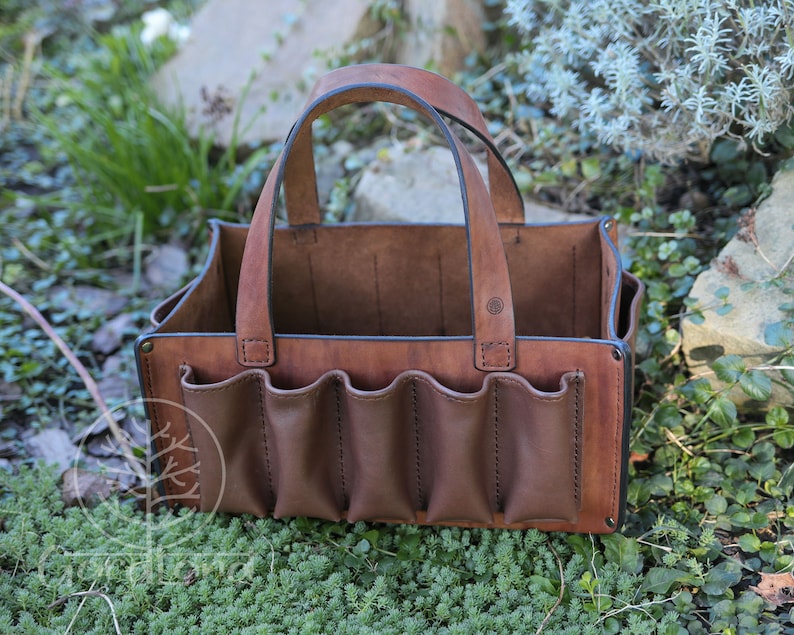 May include: A brown leather tool bag with a handle and multiple pockets. The bag is sitting on a bed of green moss.