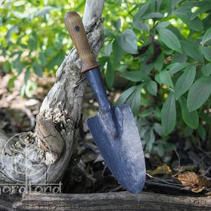 May include: A small, black gardening trowel with a wooden handle. The trowel is resting on a piece of driftwood.