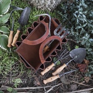 May include: A brown leather gardening tool bag with six pockets, each holding a different gardening tool. The tools include a trowel, a hand rake, a cultivator, a hand fork, and two hoes. The bag is sitting on a bed of green leaves and brown twigs.