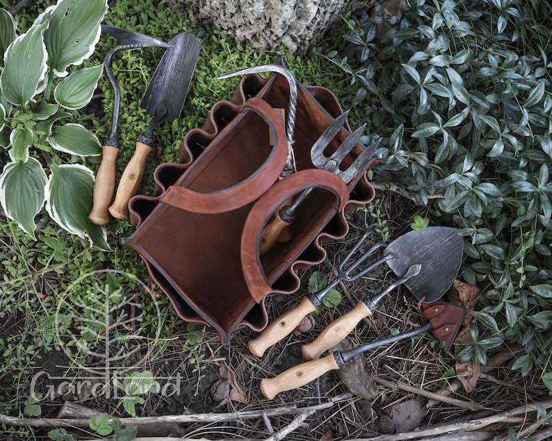 May include: A brown leather gardening tool bag with six pockets, each holding a different gardening tool. The tools include a trowel, a hand rake, a cultivator, a hand fork, and two hoes. The bag is sitting on a bed of green leaves and brown twigs.