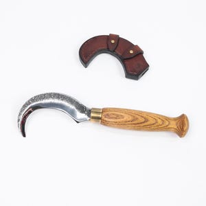 Puede incluir: Un cuchillo de hoja curva con mango de madera y funda de cuero. La hoja es plateada y el mango es de color marr&oacute;n claro con un dise&ntilde;o de vetas de madera. La funda es de color marr&oacute;n oscuro y tiene un cierre a presi&oacute;n.
