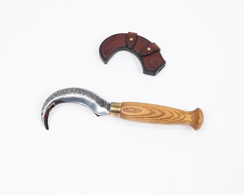 Puede incluir: Un cuchillo de hoja curva con mango de madera y funda de cuero. La hoja es plateada y el mango es de color marr&oacute;n claro con un dise&ntilde;o de vetas de madera. La funda es de color marr&oacute;n oscuro y tiene un cierre a presi&oacute;n.